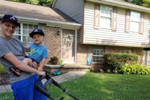 I am holding my son with one hand and a push-mower in the other hand. We are in front of our house.