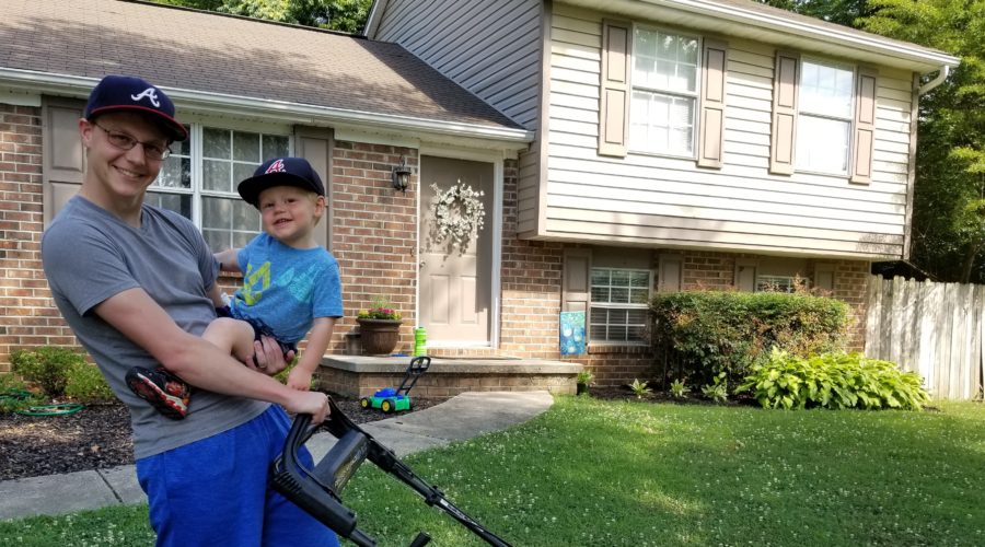 I am holding my son with one hand and a push-mower in the other hand. We are in front of our house.
